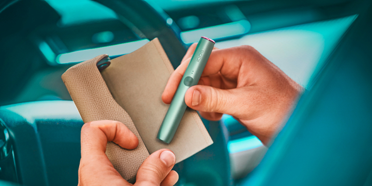 An IQOS device held by two hands in front of a car steering wheel.